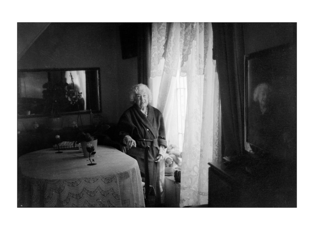 Mujer mayor junto a una ventana en el interior de una casa, en blanco y negro.
