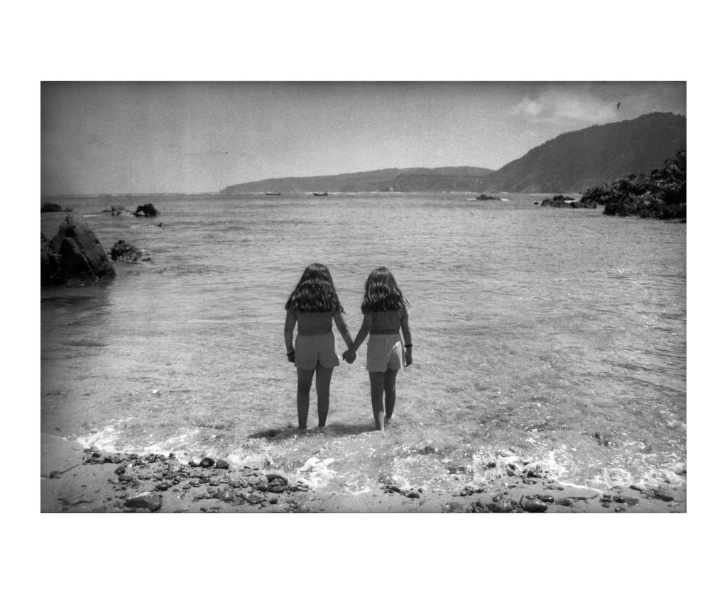 Dos niñas tomadas de la mano frente al mar, en blanco y negro.